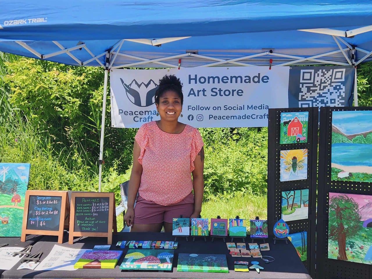 The artist standing and smiling behind her vendor booth table at Vermont Future Fest, under a blue tent with a Homemade Art Store banner, surrounded by original paintings, small art pieces, and chalkboard price signs.
