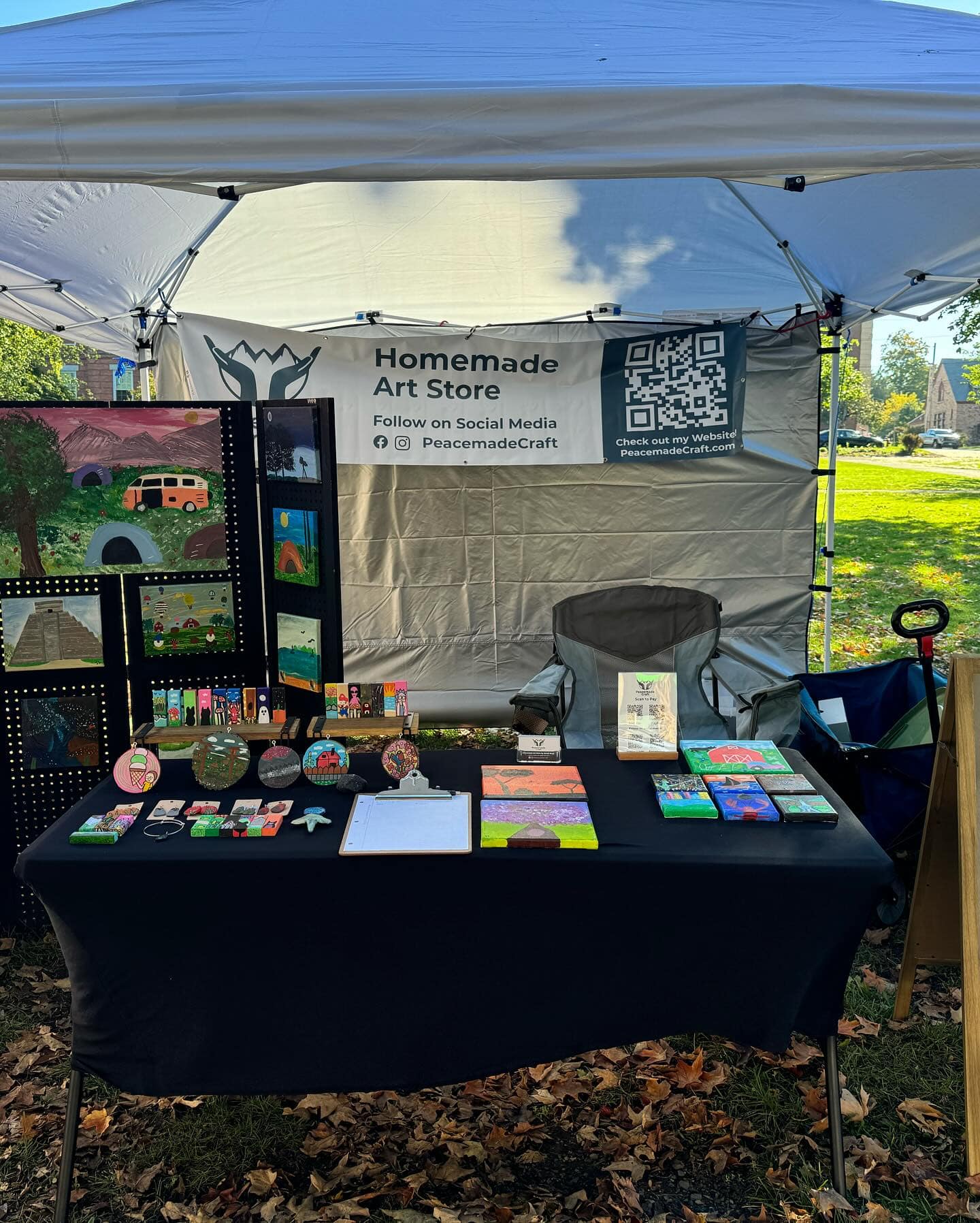 Outdoor vendor tent booth with a Homemade Art Store banner, displaying colorful acrylic paintings and handmade items on a black-clothed table.