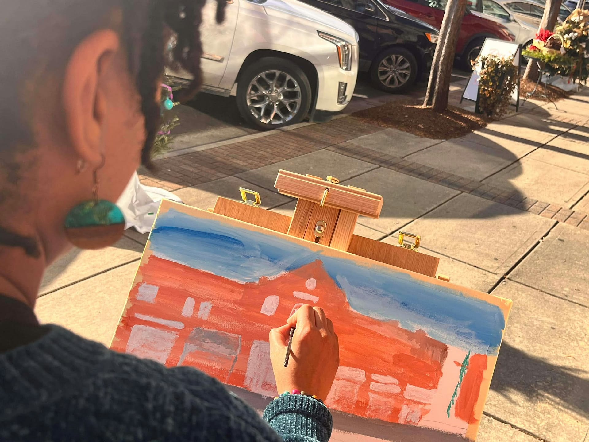 Close-up of the artist's hand painting live on an easel outdoors, working on an acrylic streetscape of a red-orange building under a blue sky.