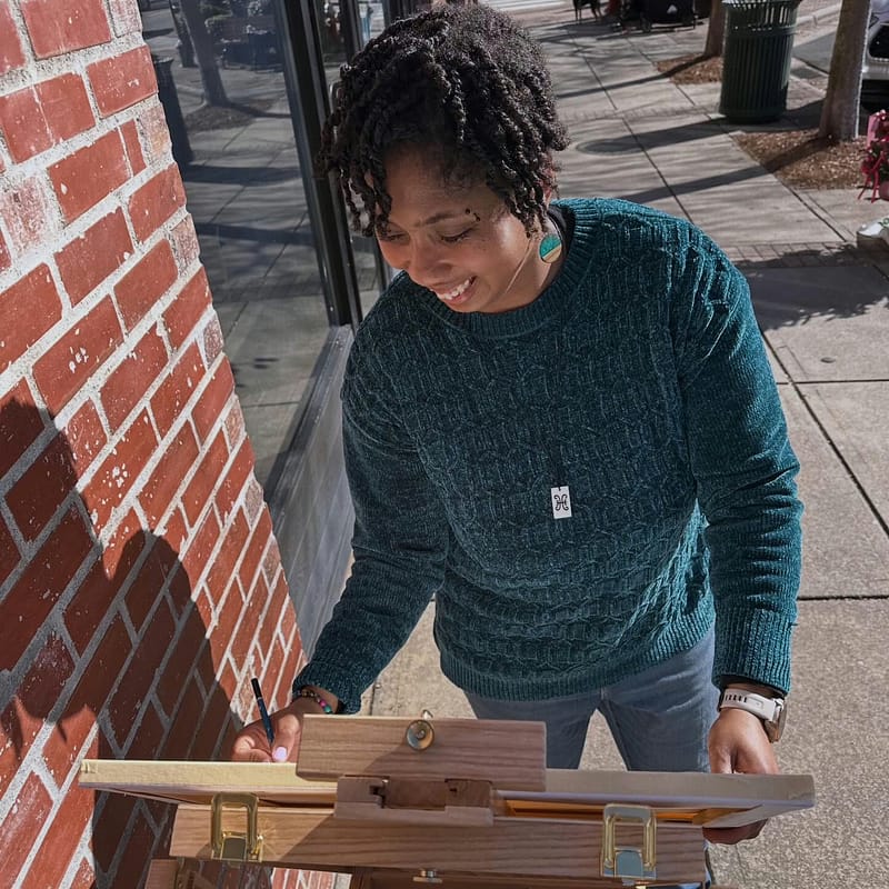 Jewell Shaw, the artist behind Peacemade Craft, painting outdoors on a portable easel near a brick storefront in Raleigh — track custom pieces like this in your My Account order history.