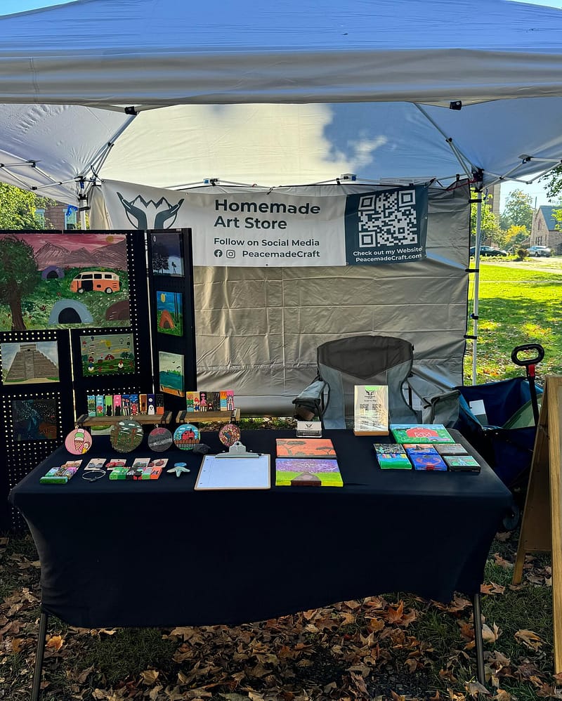 Outdoor vendor tent booth with a Homemade Art Store banner, displaying colorful acrylic paintings and handmade items on a black-clothed table.