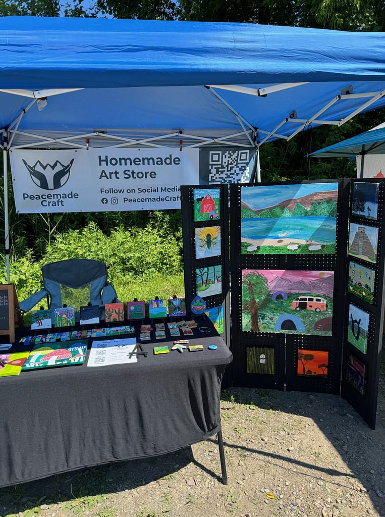 A folding table display of small original paintings and pegboard-mounted landscape artwork at an outdoor vendor booth.