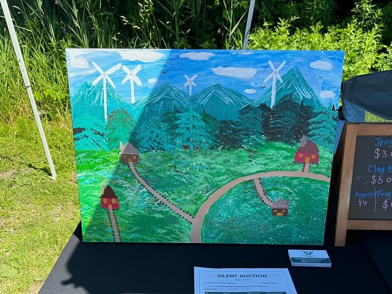 A colorful acrylic painting of a mountain landscape with wind turbines and a winding path connecting small red-roofed buildings, displayed on an easel at the vendor booth.