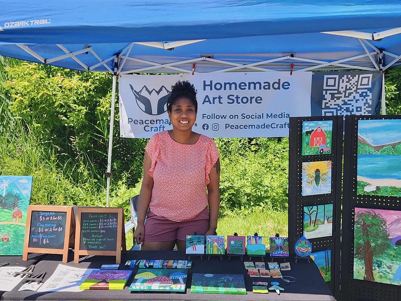 The artist standing and smiling behind her vendor booth table at Vermont Future Fest, under a blue tent with a Homemade Art Store banner, surrounded by original paintings, small art pieces, and chalkboard price signs.